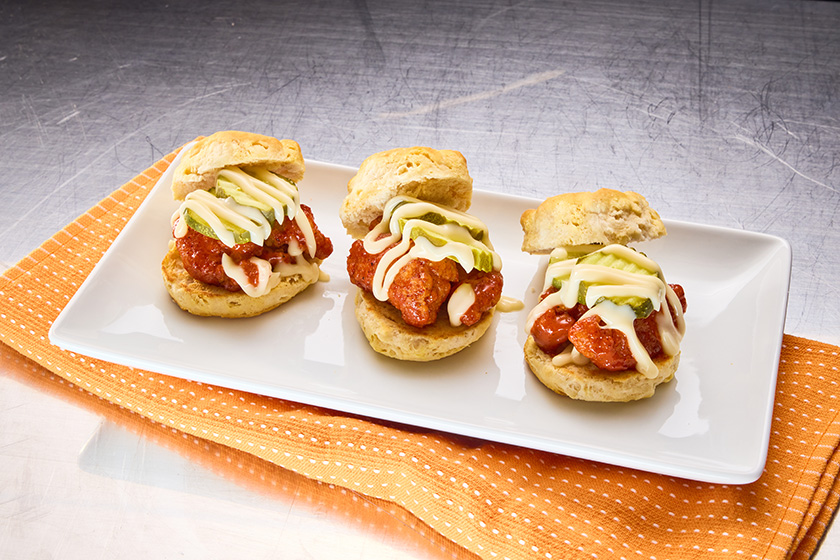 Three Nashville hot chicken biscuit sliders topped with avocado slices and Monterey Jack cheese sauce on a white platter.