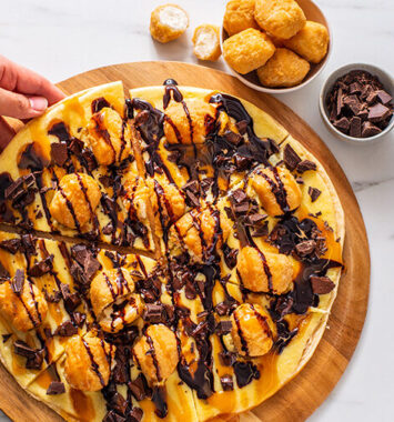 Choco-Caramel Cheesecake Pizza topped with chocolate pudding, caramel drizzle, chocolate chunks and cheesecake bites on a sliced dessert pizza crust.