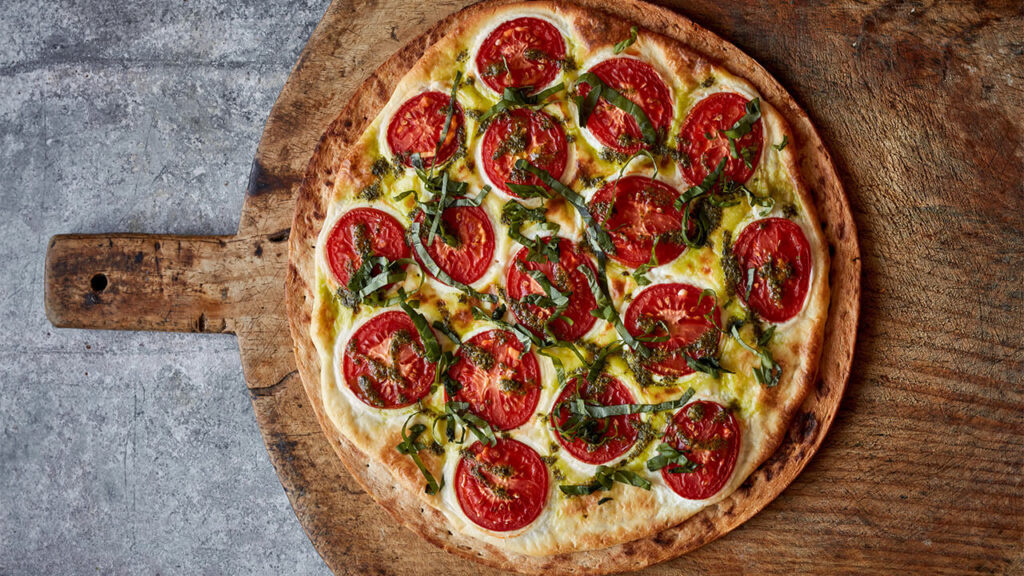 Tangy Margherita pizza topped with tomato slices, basil and green sauce drizzle on a wooden pizza peel.