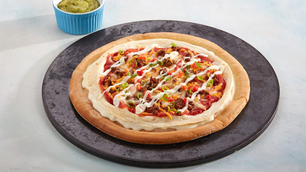 Mexican-style cheese burst pizza with layered tortilla, seasoned meat, peppers, cheese sauce drizzle and green onion on a black serving plate.