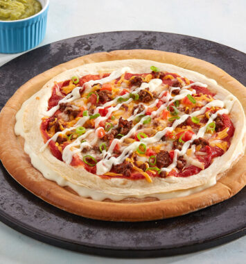 Mexican-style cheese burst pizza with layered tortilla, seasoned meat, peppers, cheese sauce drizzle and green onion on a black serving plate.